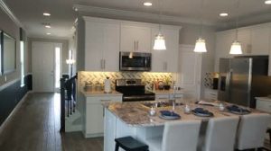 Modern kitchen with white cabinets, stainless steel appliances, a center island with granite countertop, barstools, pendant lights, and chevron tile backsplash. Listed by Rich Noto Realtor, with a hallway and front door visible in the background.