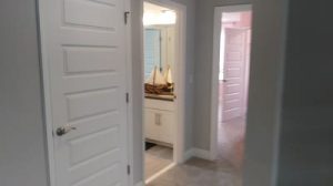 A hallway with light gray walls and white doors, one slightly open revealing a bathroom vanity with a decorative sailboat on the counter, and another door leading to a pinkish room—offered by Rich Noto Realtor.