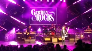 A rock band performs on stage under purple lights, with a large Garden Rocks sign displayed behind them. Four musicians play guitar, drums, and keyboard as the audience—including Rich Noto Realtor—watches from the front rows.