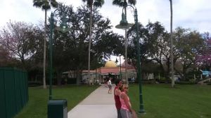 Two women stand on a wide sidewalk lined with palm trees and lampposts in a park. In the background, a yellow-domed building rises above green lawns—an inviting scene captured by Rich Noto Realtor.